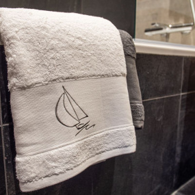 Gros plan d'un drap de douche blanc brodé d'un motif de voilier, posé sur le rebord d'une baignoire.