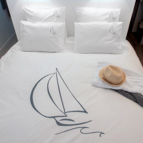 Funda de almohada blanca bordada con un velero gris y su funda nórdica a juego sobre una cama.