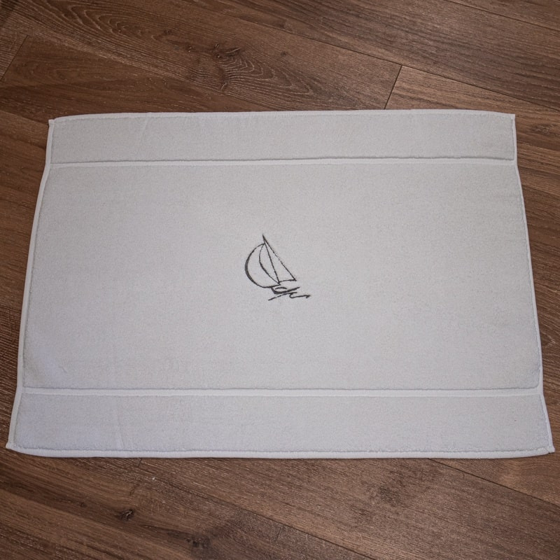 Alfombra de baño de estilo marinero, de color blanco y bordada con un motivo de velero gris.