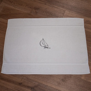 Alfombra de baño de estilo marinero, de color blanco y bordada con un motivo de velero gris.
