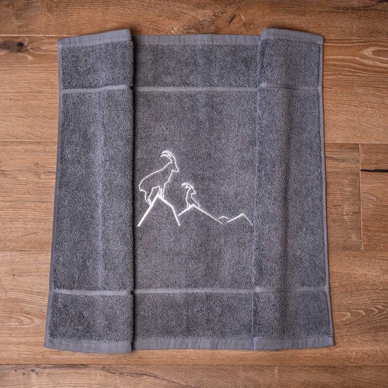 Alfombra de baño gris con motivos de montaña, bordada con un ibice blanco.