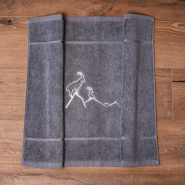 Alfombra de baño gris con motivos de montaña, bordada con un ibice blanco.