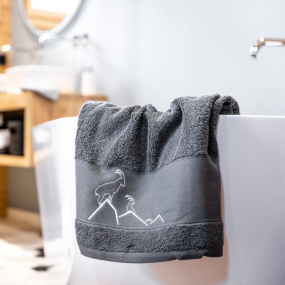 Toalla de ducha gris con motivos de montaña, con un íbice bordado en el borde de una bañera.