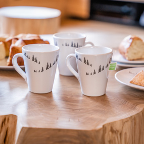 Tre mug in stile montagna di colore bianco con motivi di abeti grigi su un tavolo in legno.