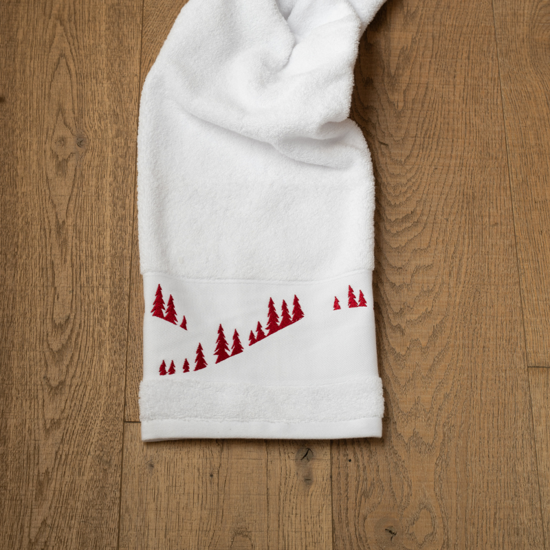 Toalla de baño blanca con motivos de montaña, bordada con un diseño de bosque de abetos.
