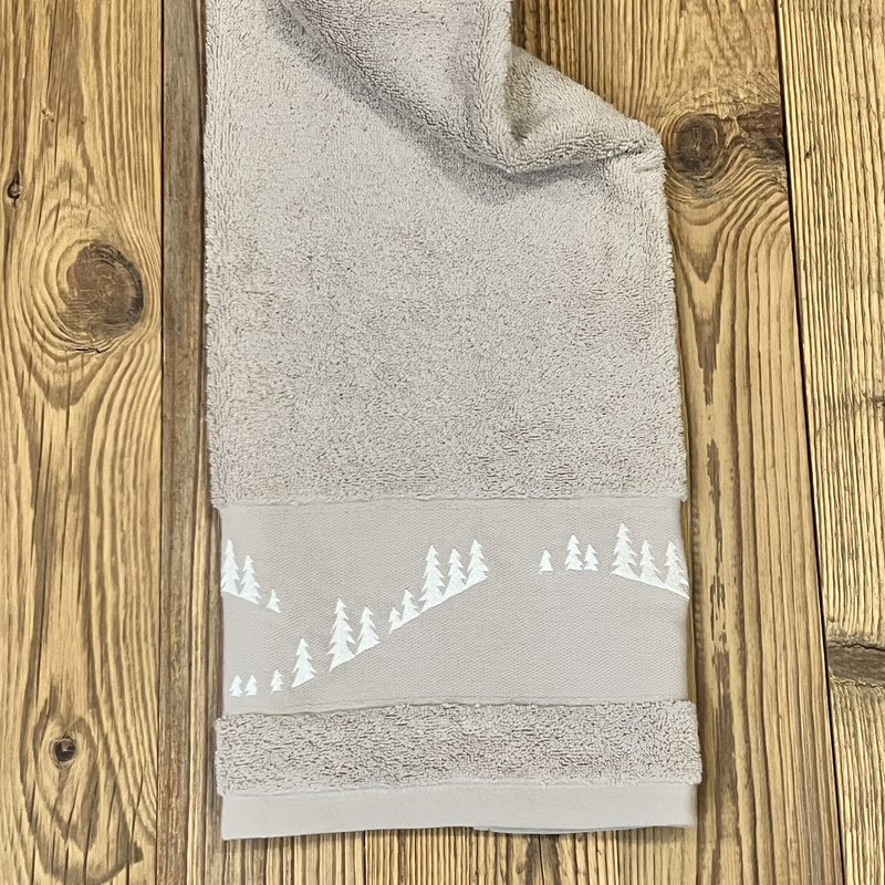 Toalla de baño con motivos de montaña en color beige, bordada con un bosque de abetos blancos.